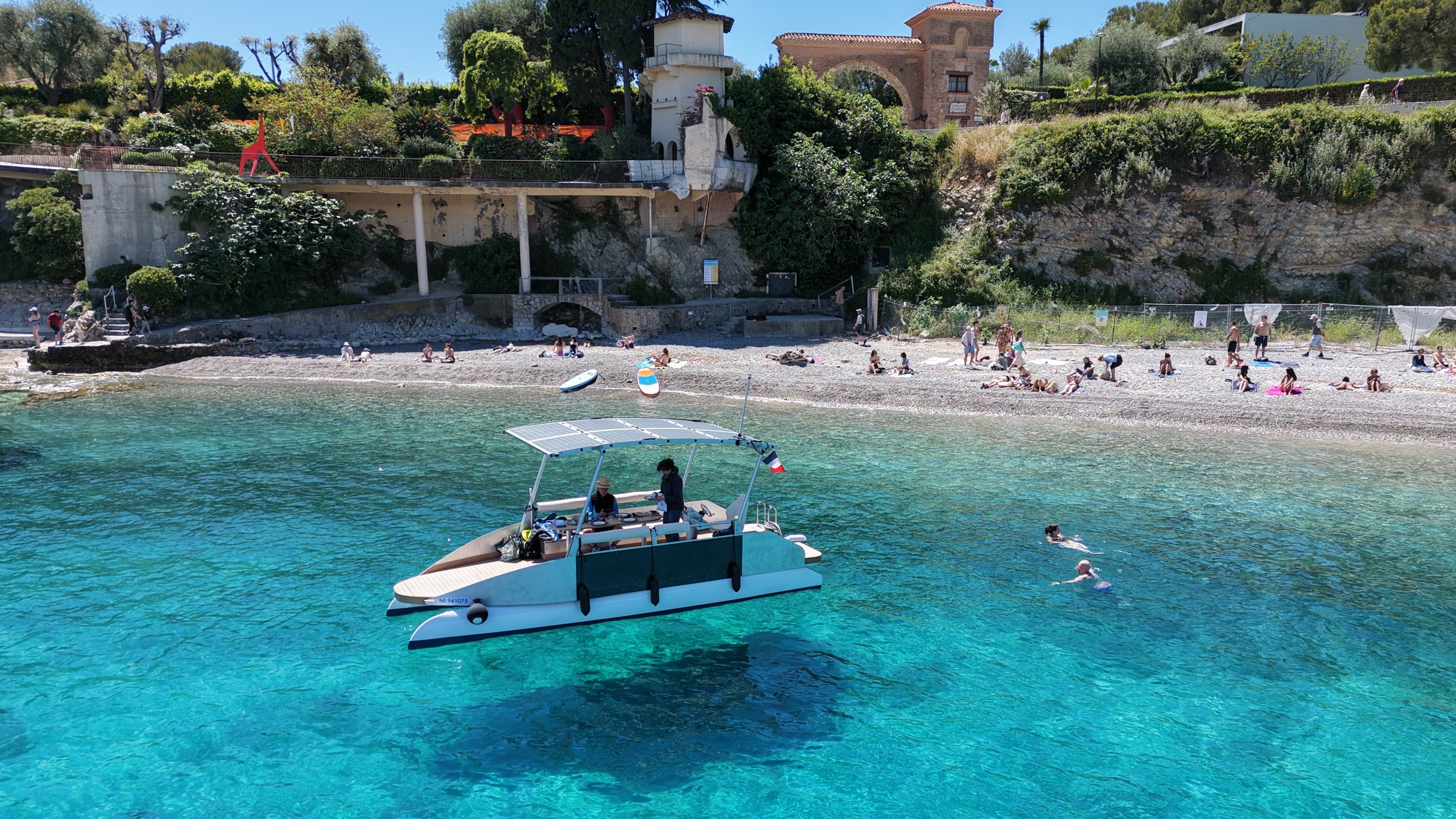 Boat with people near beach, swimmers in clear blue water, lush hillside background.