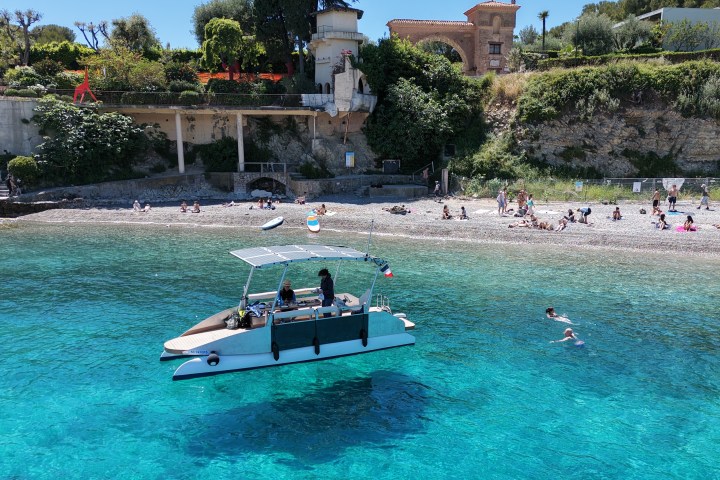 Boat with people near beach, swimmers in clear blue water, lush hillside background.