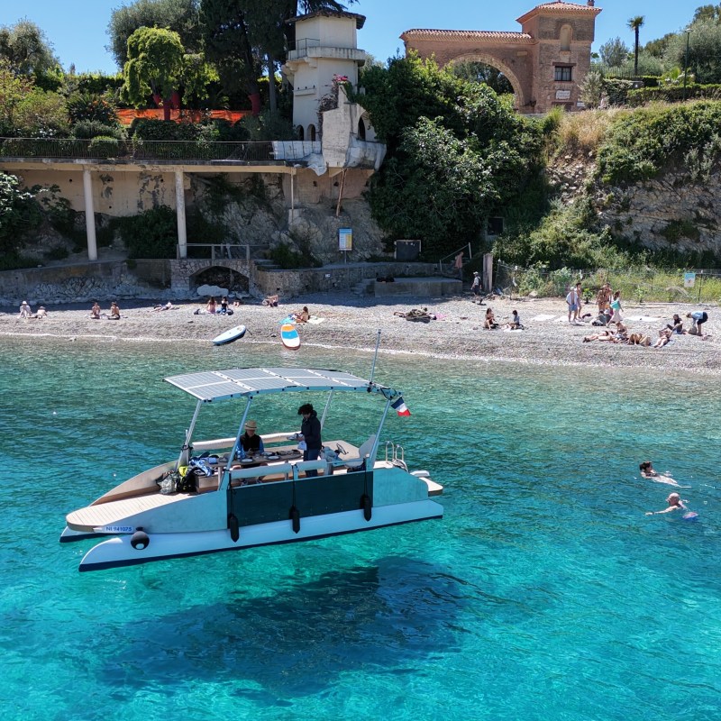 Boat with people near beach, swimmers in clear blue water, lush hillside background.