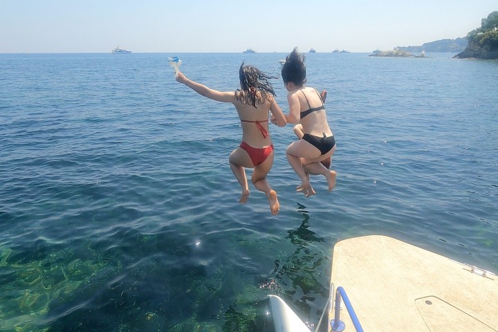 Two people in swimsuits jump off a boat into the sea, with a clear sky and distant boats visible.