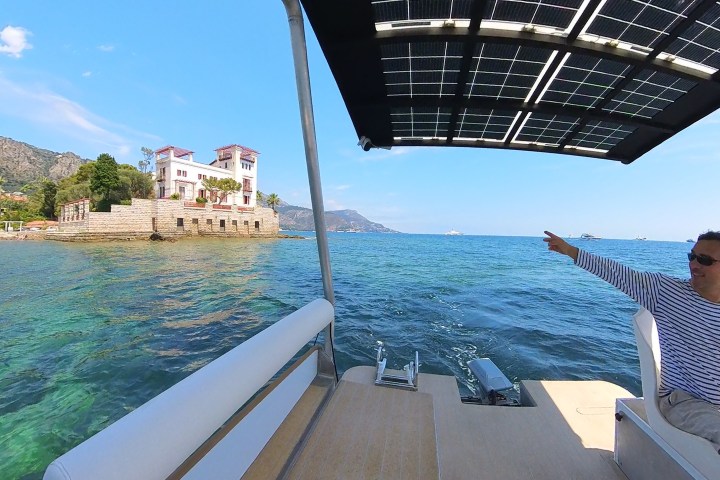Man on boat pointing towards a villa on the coast with mountains and sea in the background.
