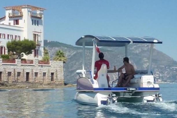 Two people on a small solar-powered boat near a coastal building.