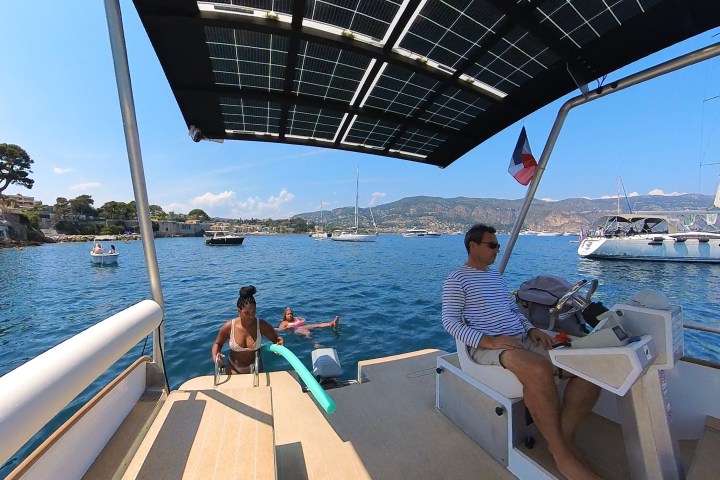 People relaxing on a boat, solar panel roof, sea view with sailboats and mountains in the background.