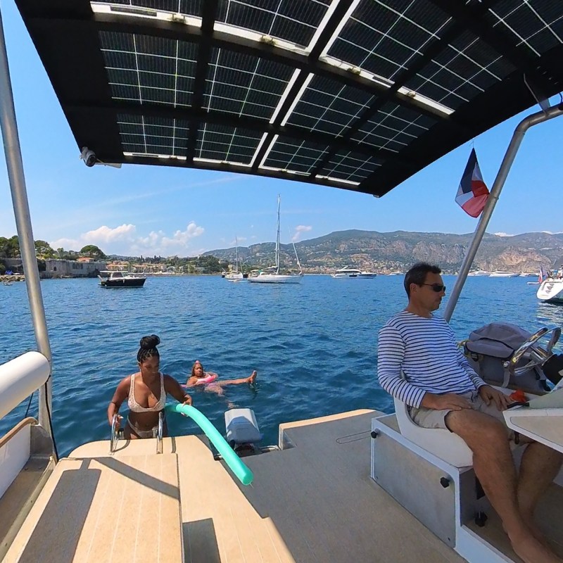 People relaxing on a boat, solar panel roof, sea view with sailboats and mountains in the background.