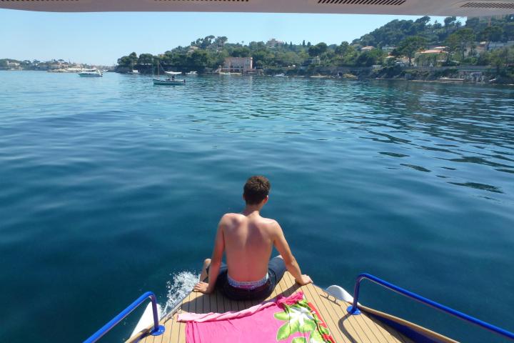 Person sitting on boat's edge with pink towel, facing calm sea and distant coastline.