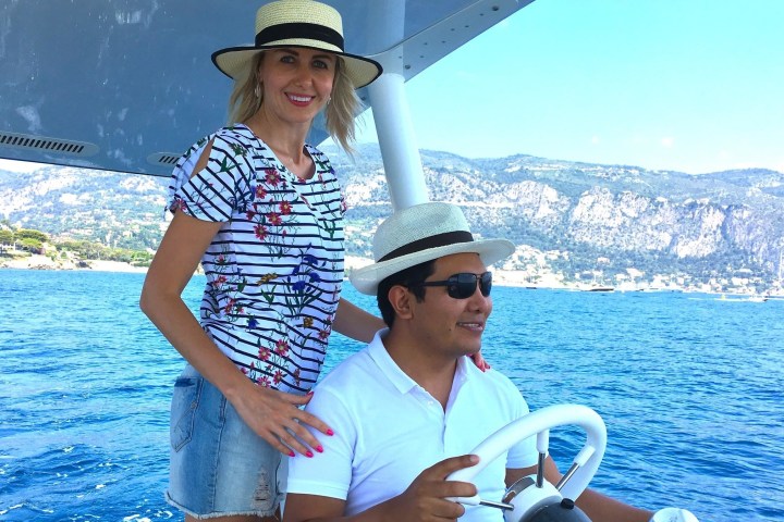 Two people in hats on a boat in front of a mountainous coastline.