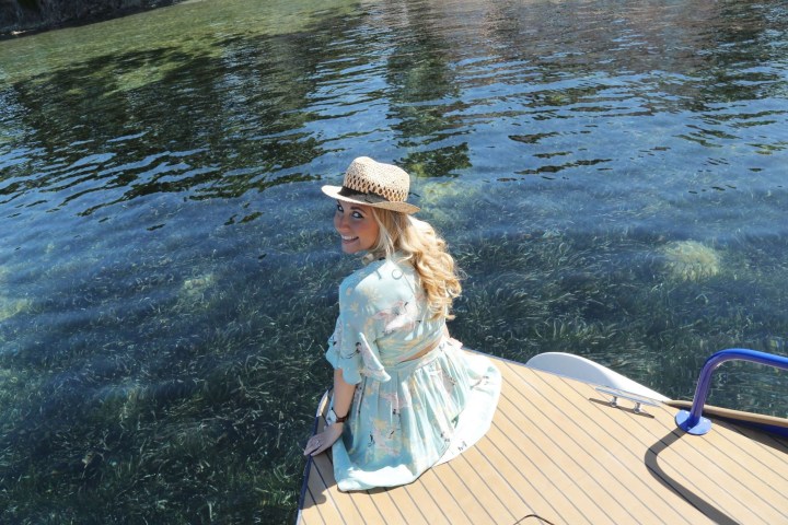 Smiling woman in hat looking back while seated on a boat near clear water.