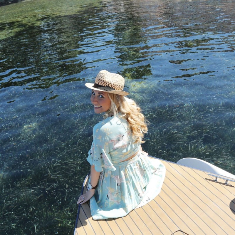 Smiling woman in hat looking back while seated on a boat near clear water.