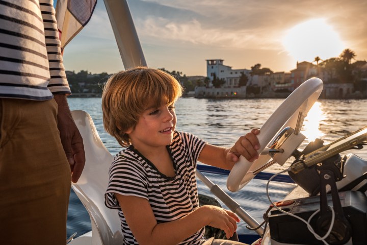Child steering a boat at sunset with an adult nearby. Water and buildings in the background.
