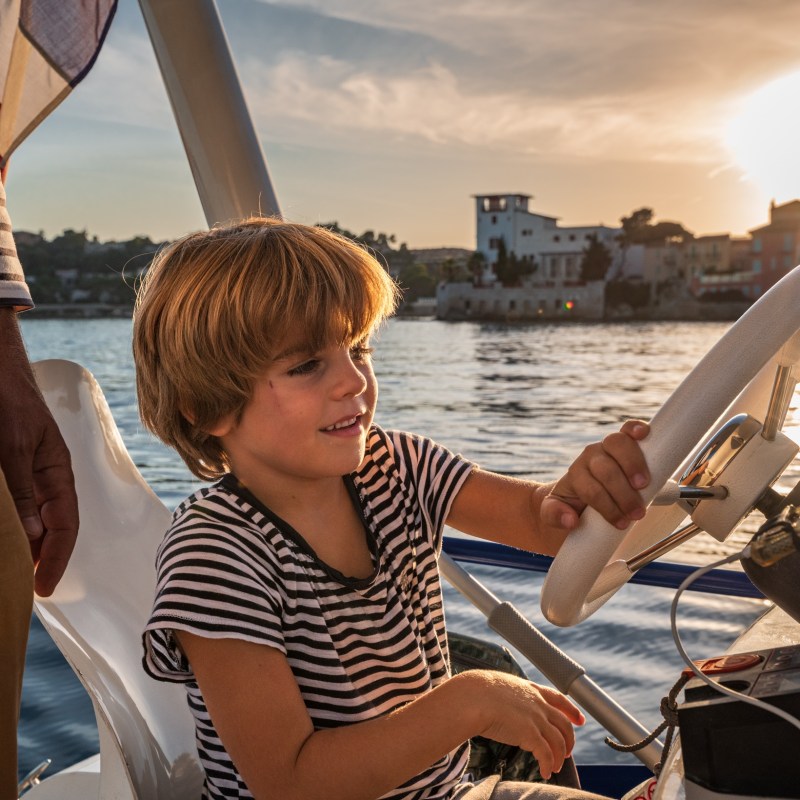 Child steering a boat at sunset with an adult nearby. Water and buildings in the background.