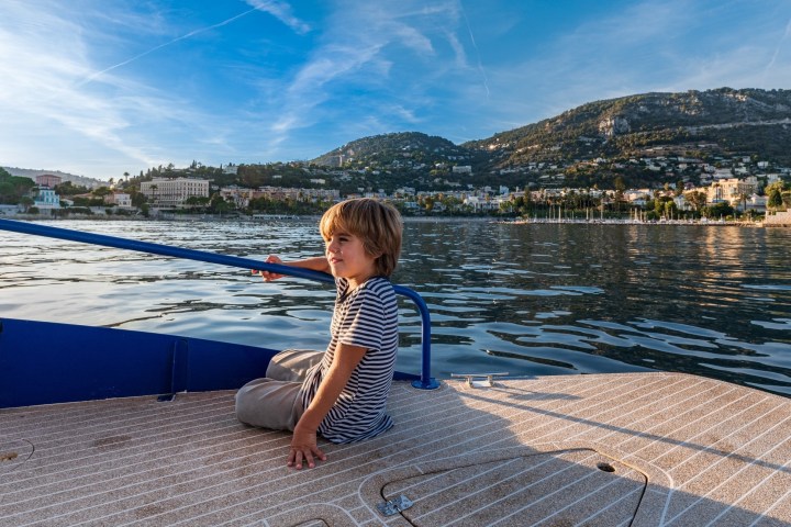 Child sitting on a boat enjoying scenic lake and hillside town view under a clear blue sky.