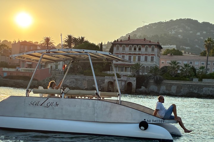 Two people relax on a boat at sunset near a coastal town with hills.