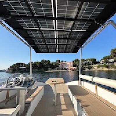 Boat with solar panel canopy on calm water, shoreline in background.