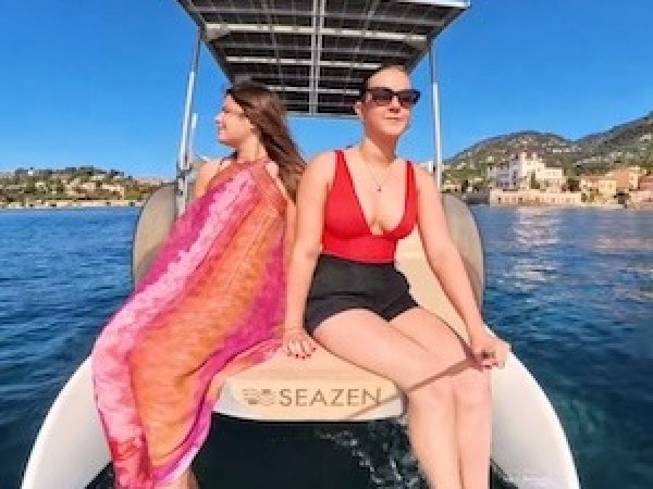 Two women in swimsuits on a boat with solar panels in a sunny coastal area.