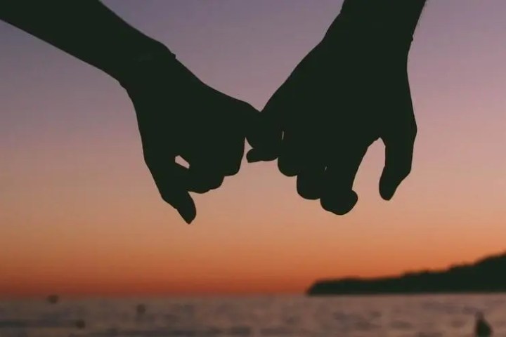 Silhouette of two hands holding pinkies at sunset over water.