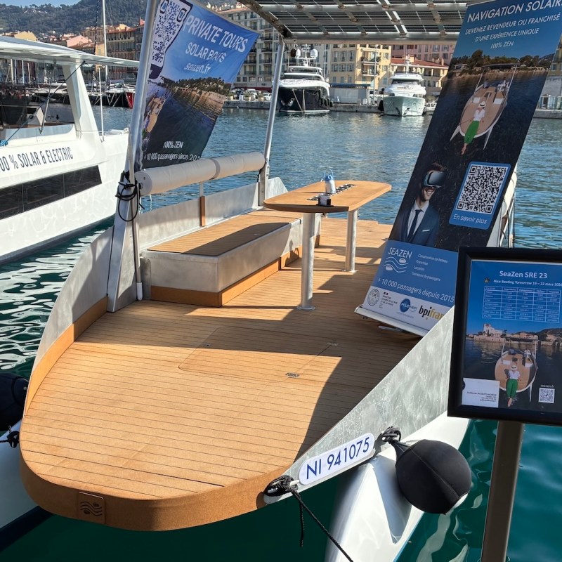 Small boat with wooden deck docked in marina with city and mountains in background.