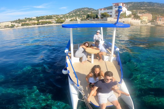 People on a small boat with a canopy, clear water, and coastal town in the background.