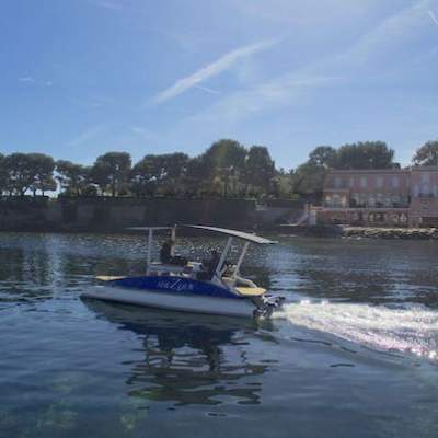 Solar-powered boat on calm water near a shoreline with trees and buildings.